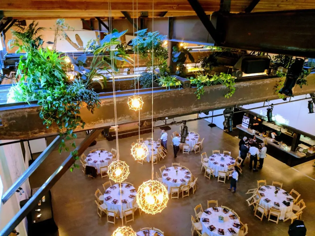 Inside The Pearl: round tables set for dinner under woven globe lights, with hanging plants along the mezzanine and a bar along the wall